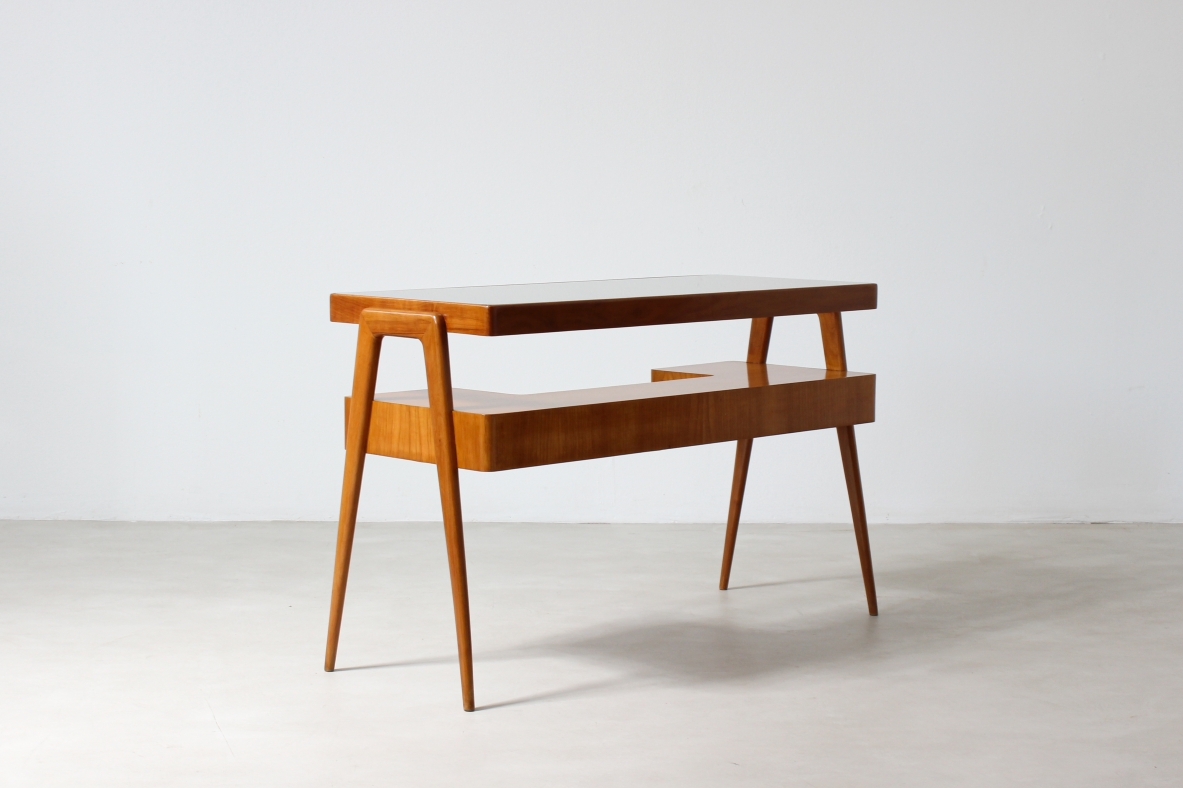 Small desk in blond wood with glass top and shaped shelf with two drawers on the front.